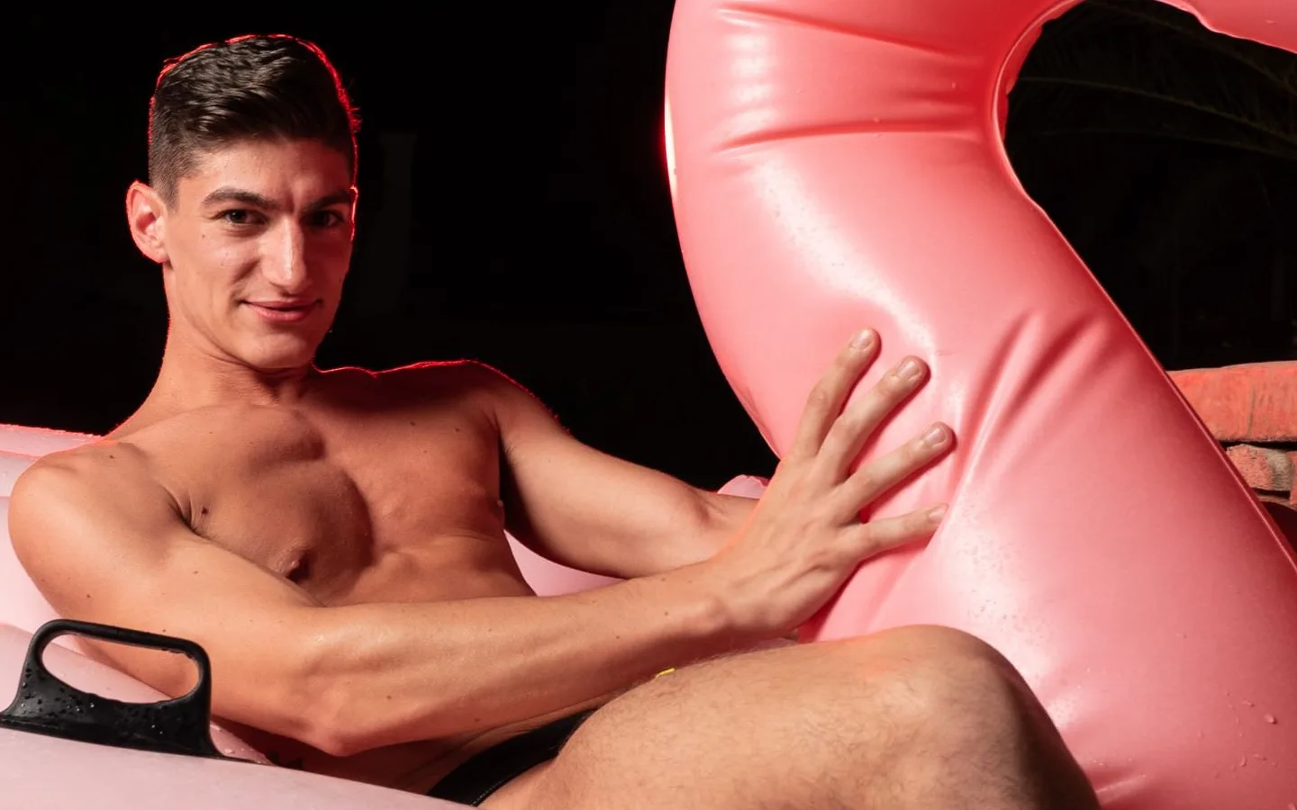 Athletic Twinks model Aiden Ward, shirtless and smiling, lounging on a pink flamingo pool float at night