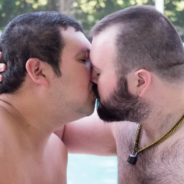 Two shirtless, hairy Bear Chubs bears kissing poolside, one cradling the other's head