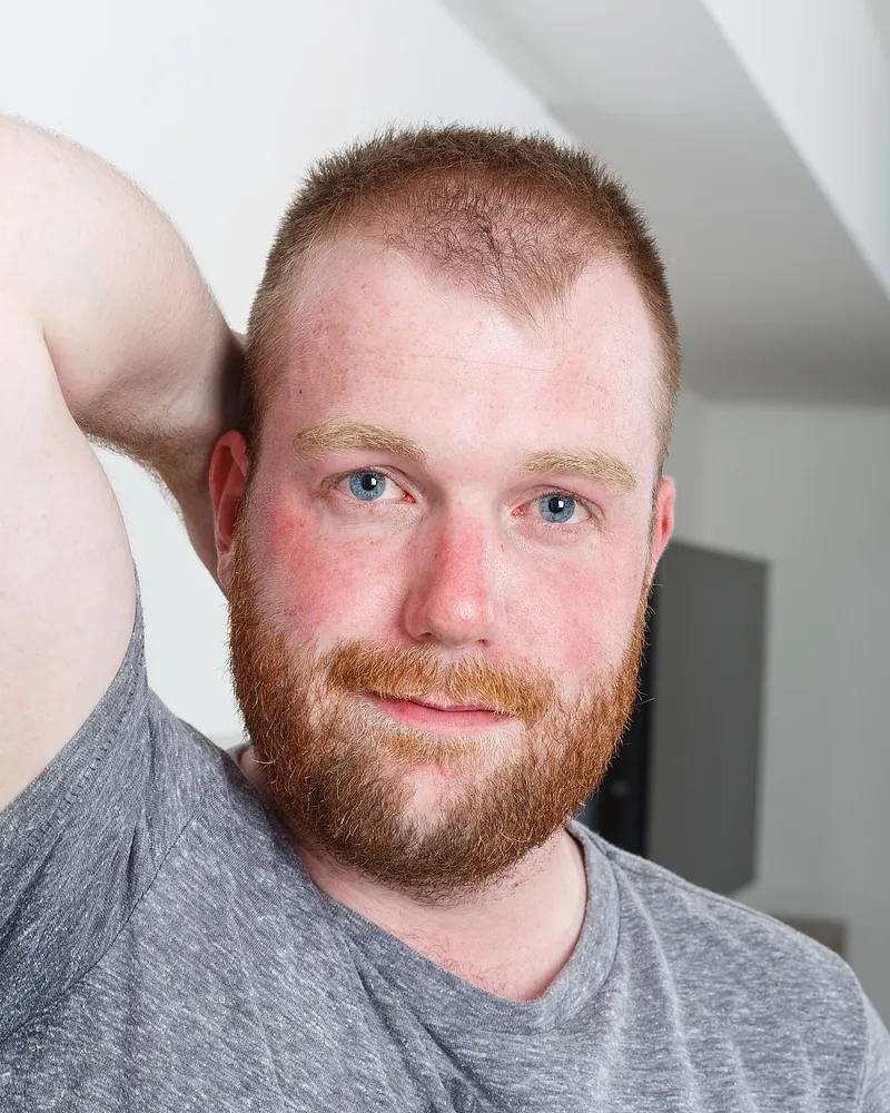 Smiling red-bearded man in a grey T-shirt with one arm raised behind his head, model portrait from Bear Films