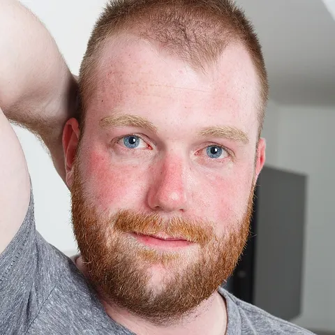 Smiling red-bearded man in a grey T-shirt with one arm raised behind his head, model portrait from Bear Films