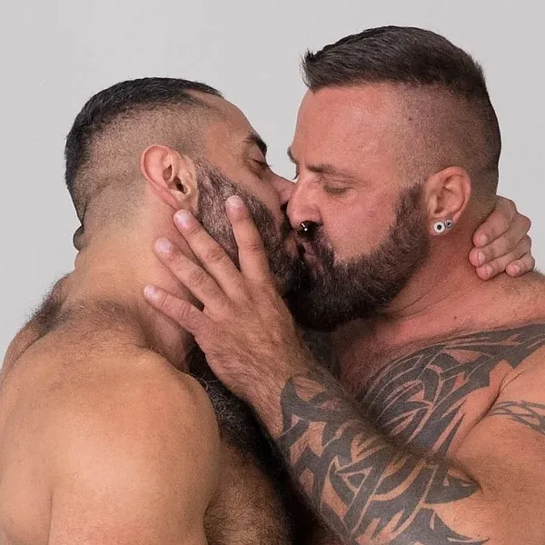 Two shirtless, tattooed, muscular Bear Films hunks kissing in a tight embrace, wearing silver shorts