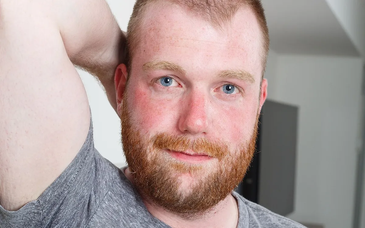 Smiling red-bearded man in a grey T-shirt with one arm raised behind his head, model portrait from Bear Films