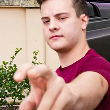 Promotional still from Gay Wire showing a man in a maroon T-shirt outdoors beside a car