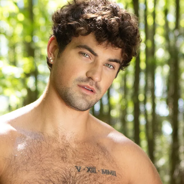 Men.com model with curly hair and a hairy chest, shirtless in a sunlit forest, wearing Men.com-branded briefs