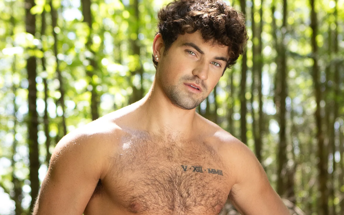 Men.com model with curly hair and a hairy chest, shirtless in a sunlit forest, wearing Men.com-branded briefs